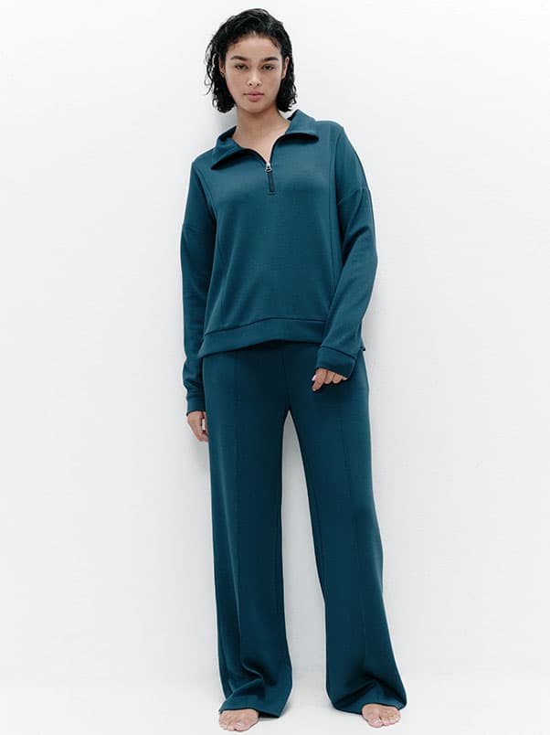 Person wearing a teal zip-up pullover and matching wide-leg pants, standing barefoot against a plain white background.