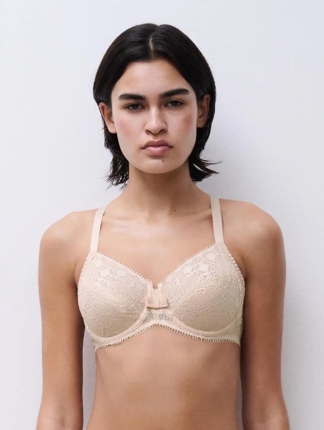 Person wearing a beige lace bra with thin straps, standing against a plain white background.