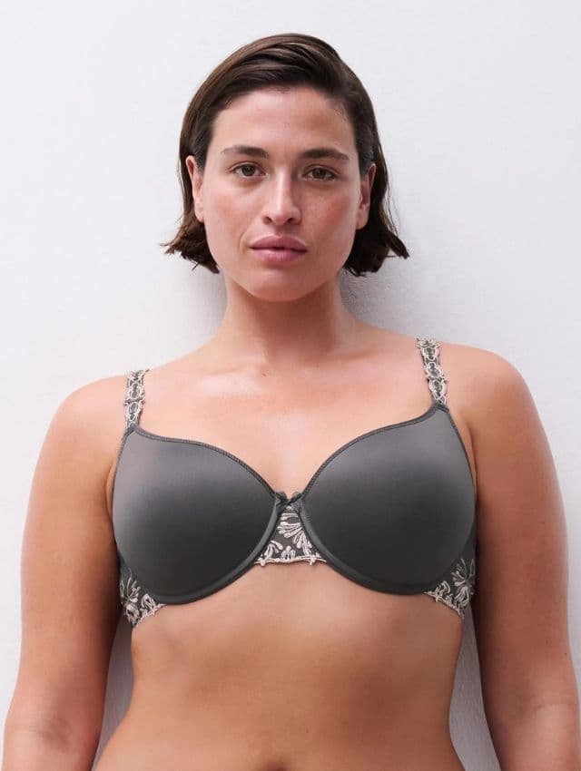 Person with short hair wearing a gray bra with patterned straps, standing against a plain white background.