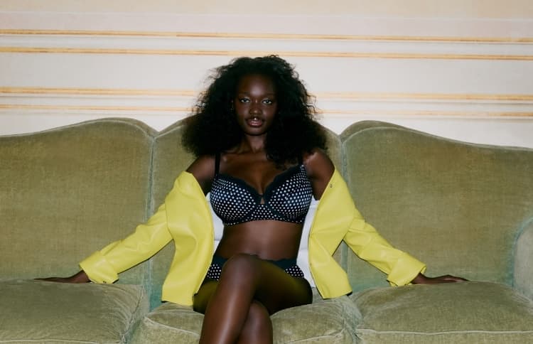 Person in a polka dot lingerie set and yellow jacket sits confidently on a green sofa with crossed legs, against a neutral background.