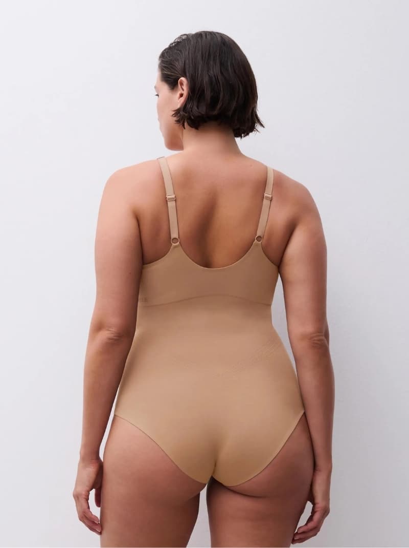 Person with short hair wearing a beige bodysuit stands facing a plain light background, showing the back design and adjustable straps.
