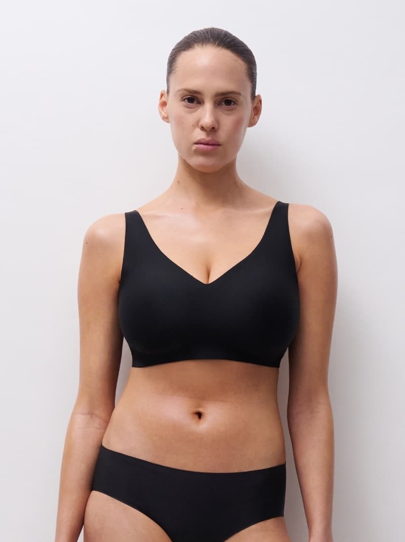 Woman in black undergarments stands against a plain white background, looking directly at the camera.