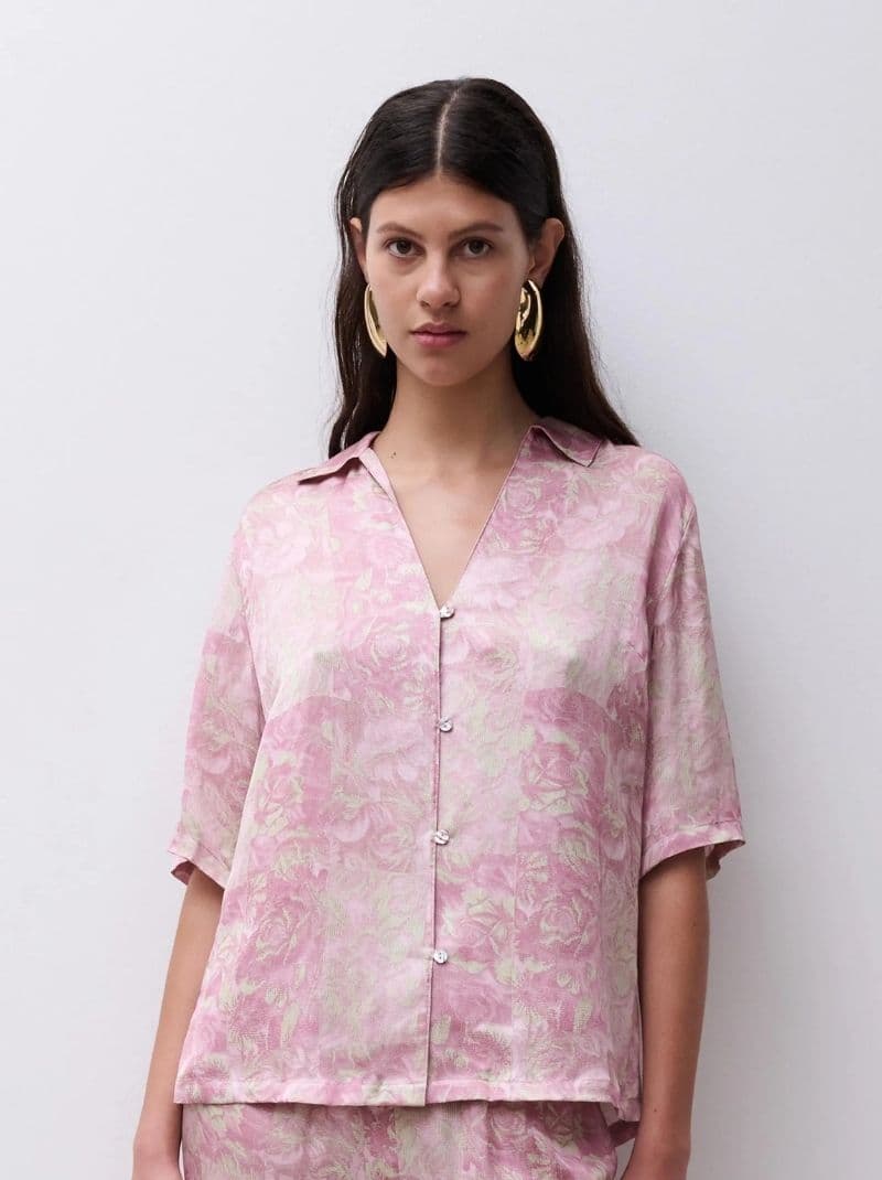 Woman wearing a pink patterned shirt with large gold hoop earrings, standing against a plain background.