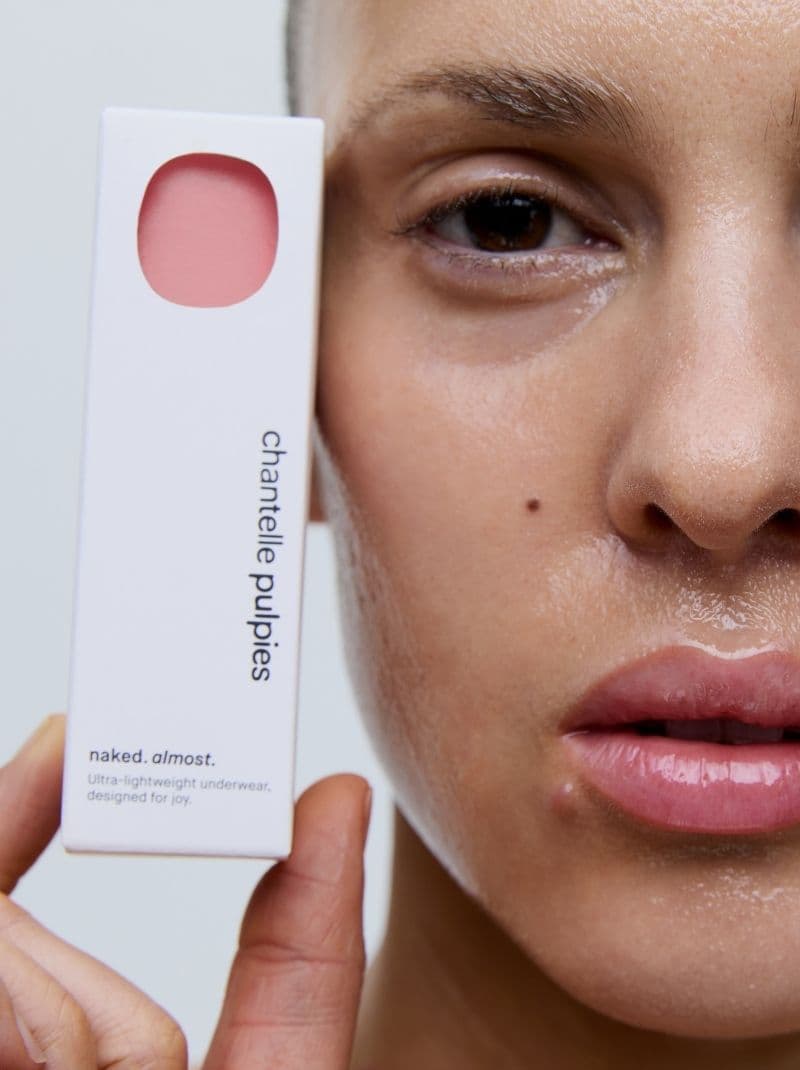 Close-up of a person's face with glossy skin holding a box labeled "chantelle puppies," featuring a cutout showing pink fabric.