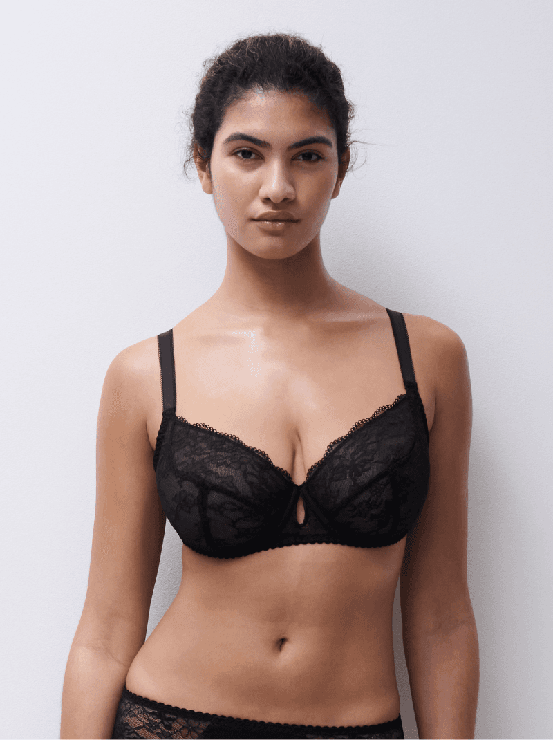 A person wearing a black lace bra stands against a white background, looking directly at the camera.