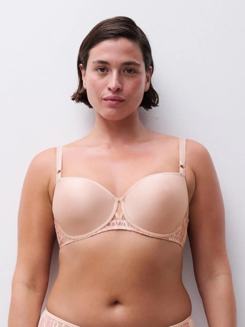 Person in a nude bra and matching underwear, standing against a white background. Short brown hair and a confident expression.