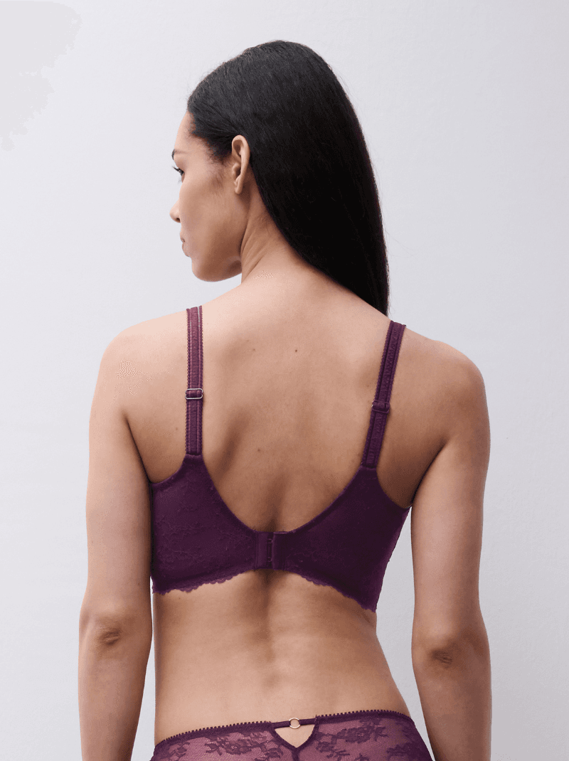 Woman with long dark hair facing away, wearing a purple lace bra and matching underwear, against a plain background.