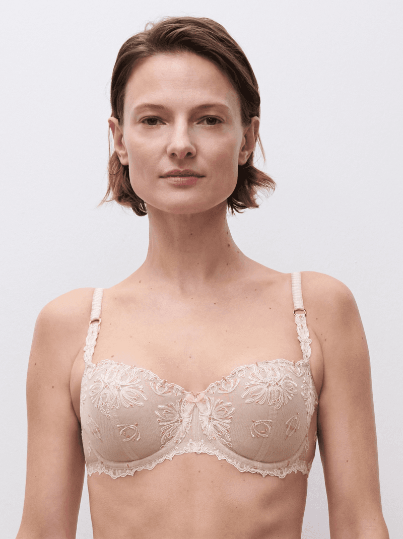A woman with short hair is wearing a beige lace bra with floral patterns, posing against a plain white background.