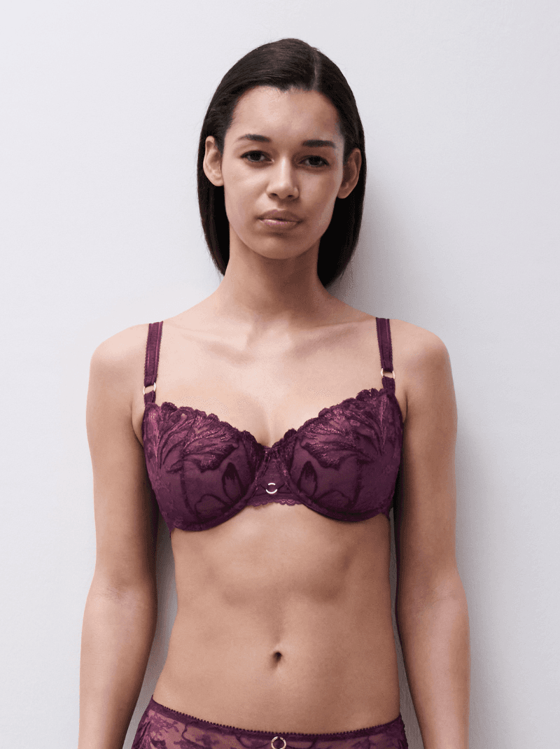 A woman wearing a purple lace bra and matching underwear stands against a light background.