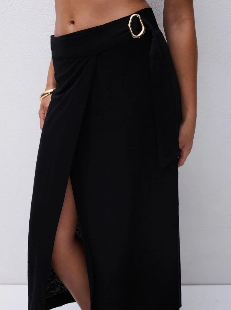 Woman wearing a high-waisted black wrap skirt with a side slit and gold buckle, against a white background.