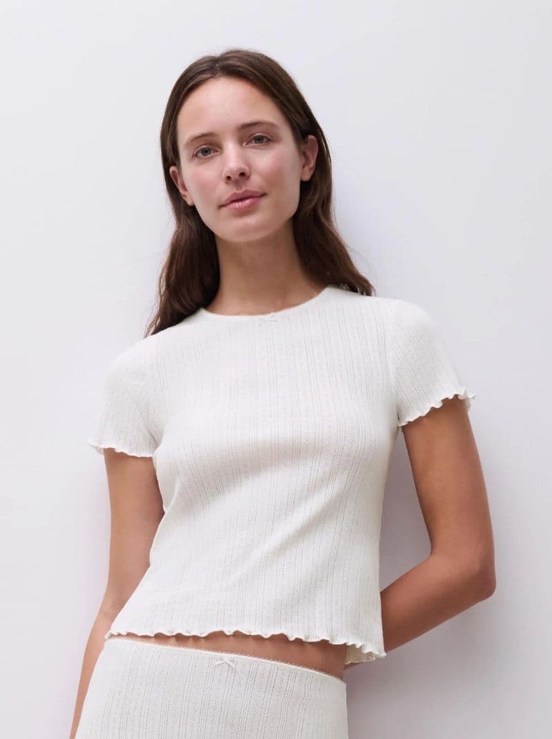 Woman in a white, short-sleeved, ribbed shirt with scalloped edges, standing against a plain white background.