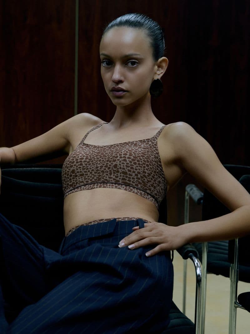 Person sitting confidently in a chair, wearing a leopard print top and pinstriped pants, with dark wooden background.