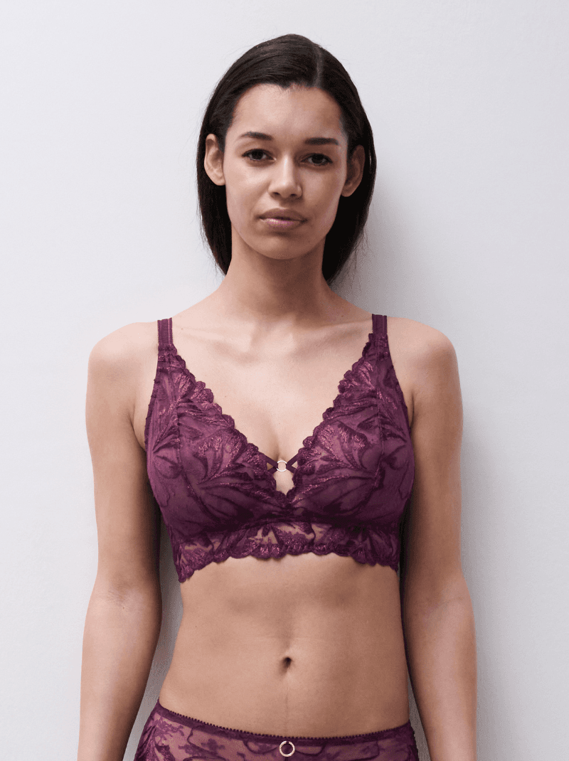 A woman wearing a purple lace bra with floral patterns stands against a plain background.