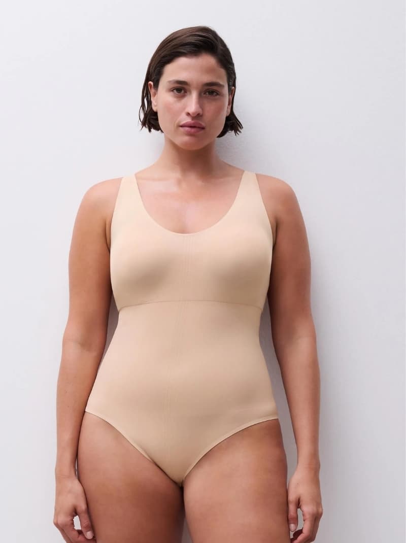A woman in a beige shapewear stands against a white background, looking directly at the camera with a neutral expression.