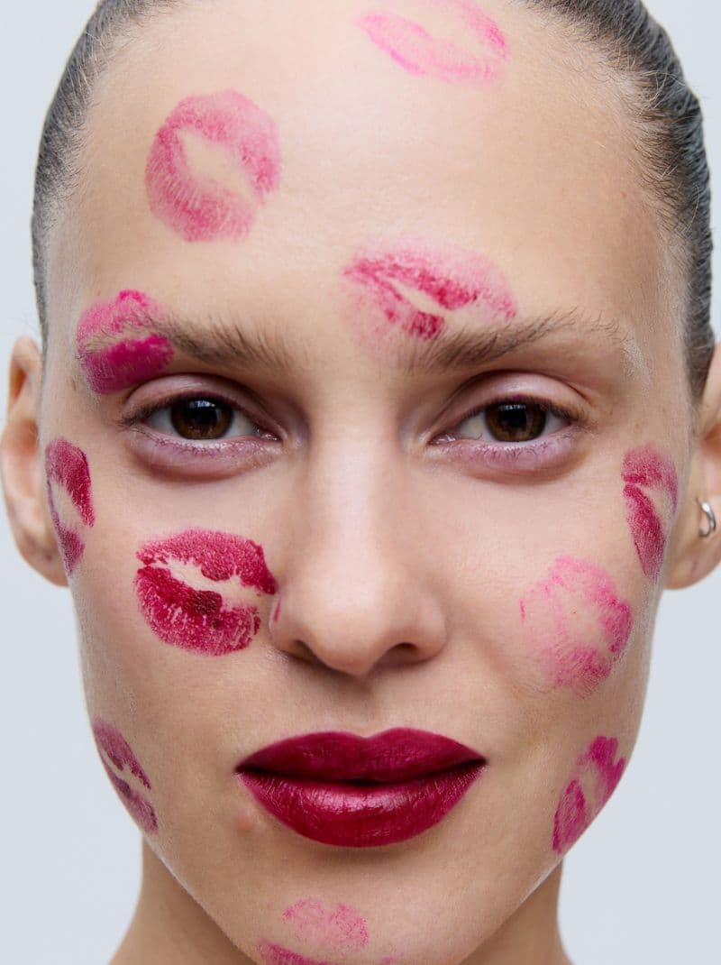 A person with multiple lipstick kiss marks on their face, wearing bold red lipstick, against a plain background.