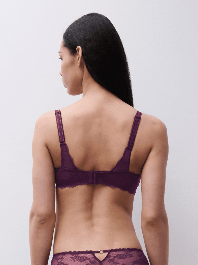 A woman with long dark hair is shown from behind wearing a purple lace bra and matching underwear, standing against a light background.