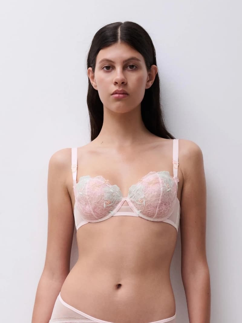 Person wearing a pastel pink lace bra with floral embroidery, standing against a plain white background.