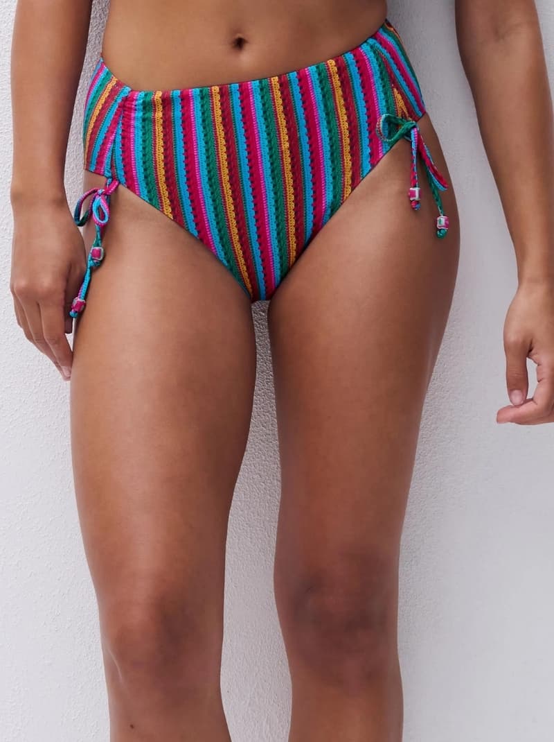 Person wearing colorful, striped, high-waisted bikini bottom with side ties, standing against a light wall.