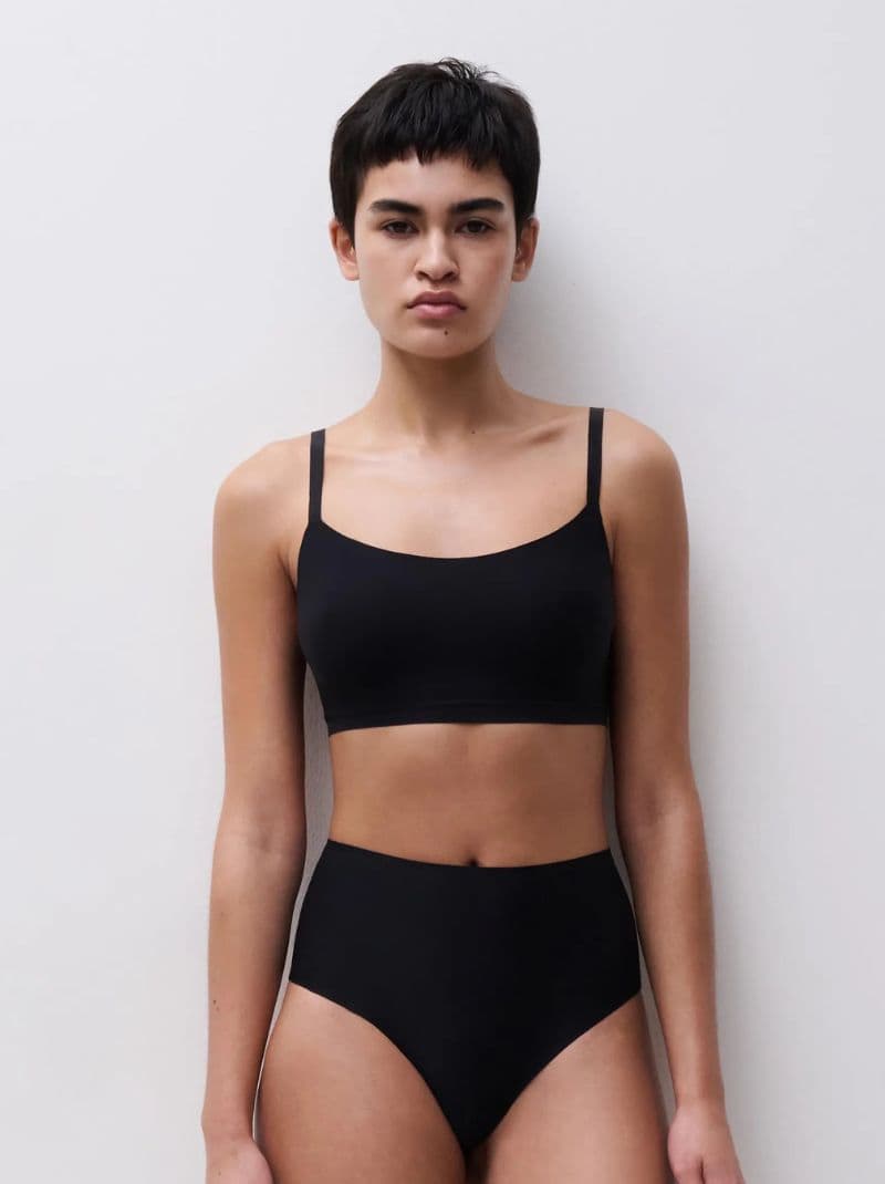 Person with short hair wearing a black bra and high-waisted underwear, standing against a plain white background.