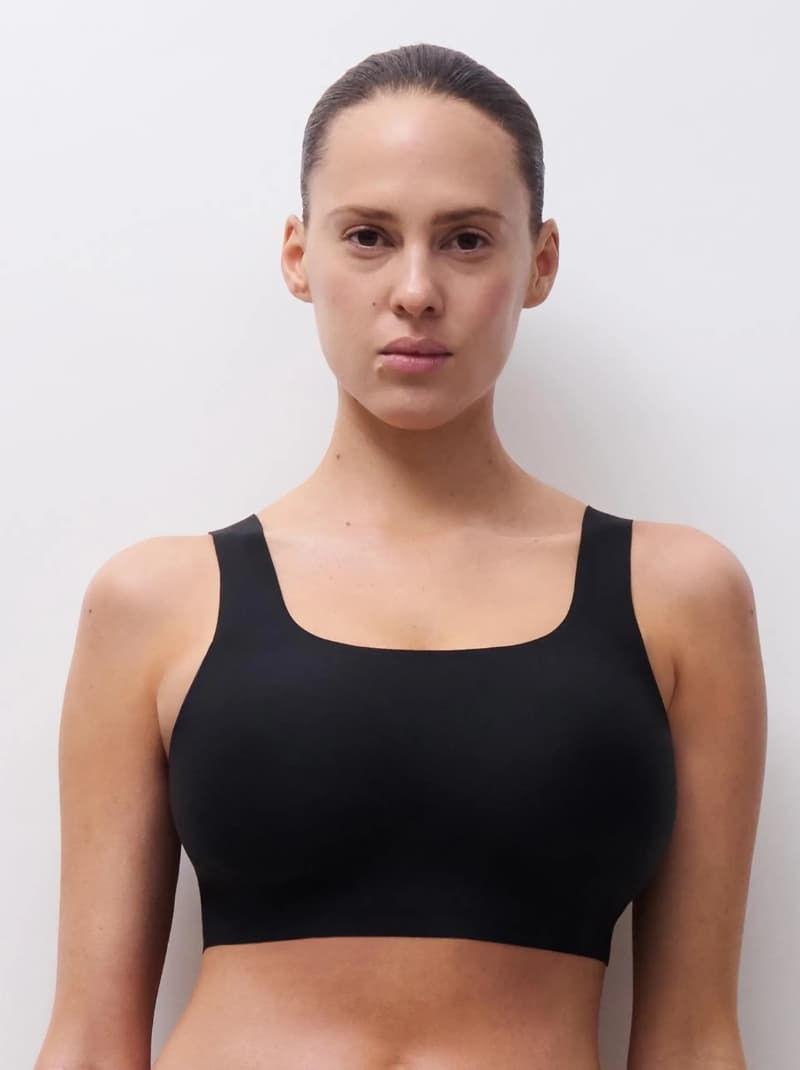 Woman in a black bra stands against a plain white background, displaying a neutral expression.