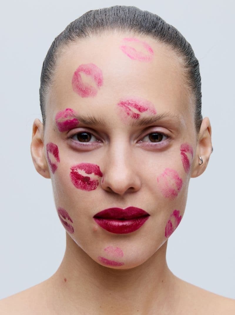 A person with slicked-back hair and bold red lips, their face marked with multiple red lipstick kiss prints.