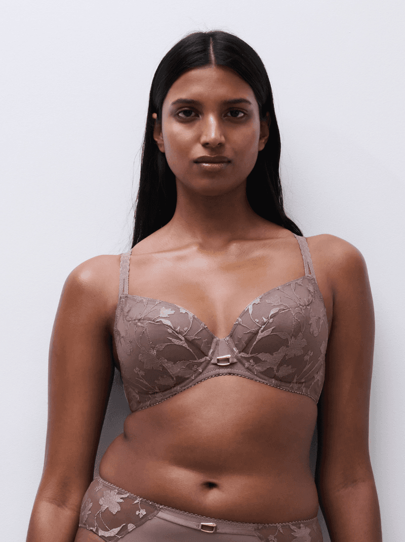 A model wearing a taupe-colored, floral lace bra and matching underwear stands against a plain white background.