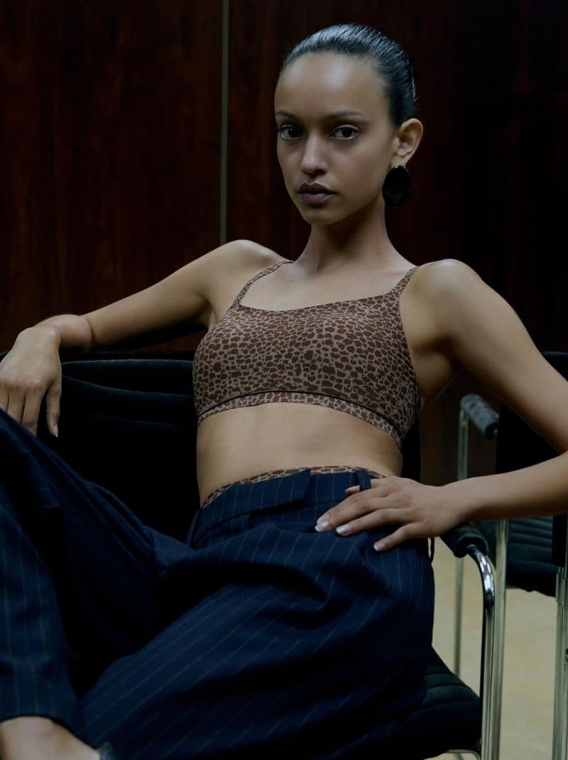 Person in a leopard print top and striped pants seated on a chair, posing confidently against a dark wooden background.