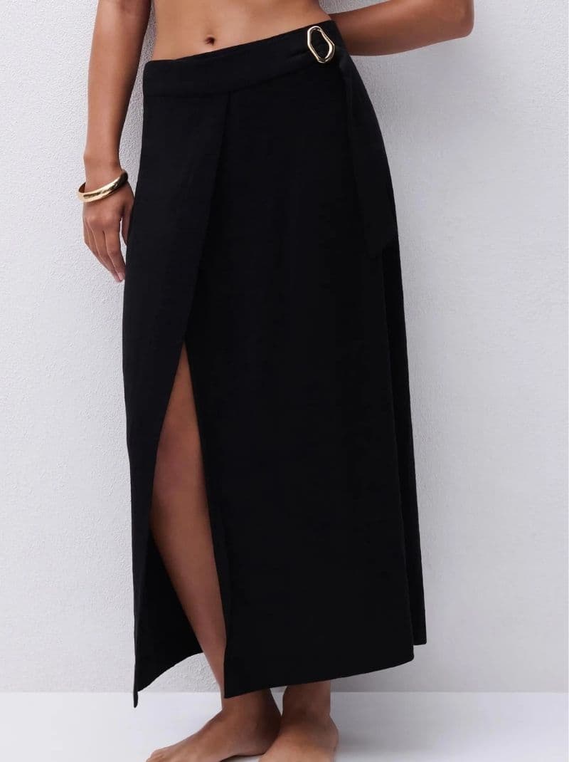 Person wearing a high-waisted black wrap skirt with a front slit and gold buckle, accessorized with a gold bracelet, against a white background.
