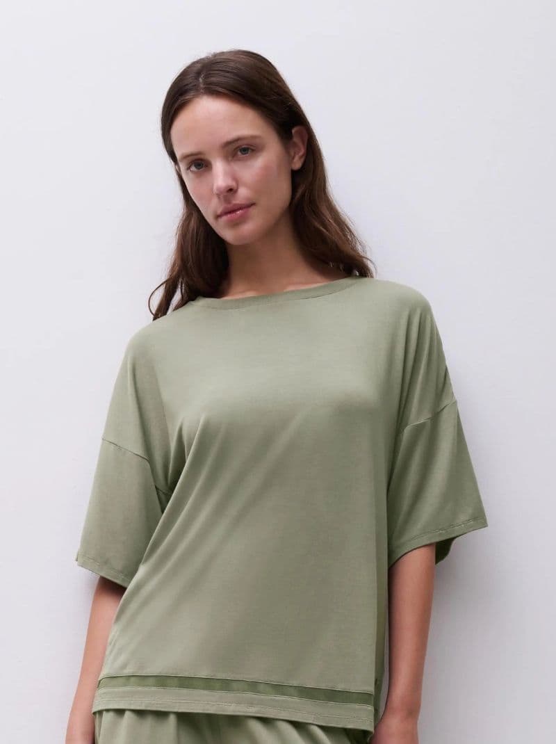 A woman in a loose green top stands against a plain white background, looking directly at the camera.