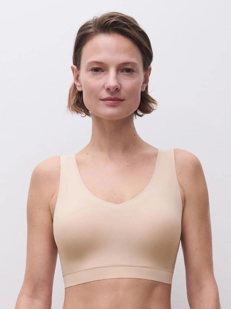 A person wearing a golden beige, sleeveless top stands against a plain, light background, looking directly at the camera.