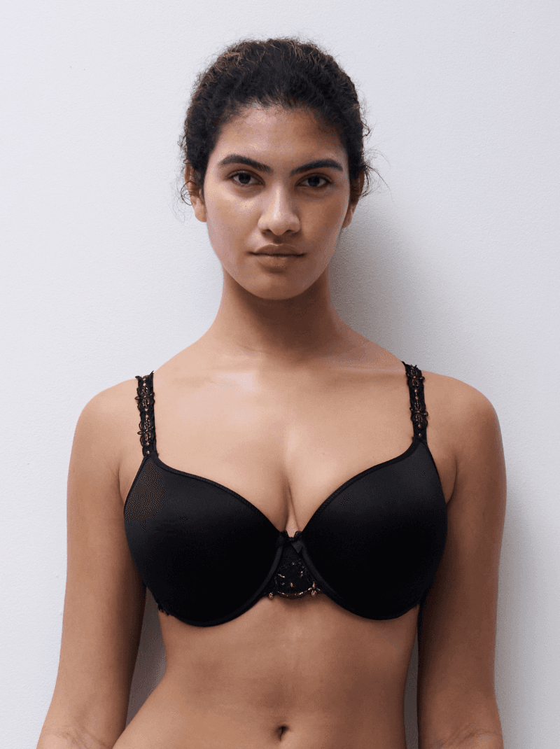 A woman wearing a black bra with lace detailing stands against a plain white background.