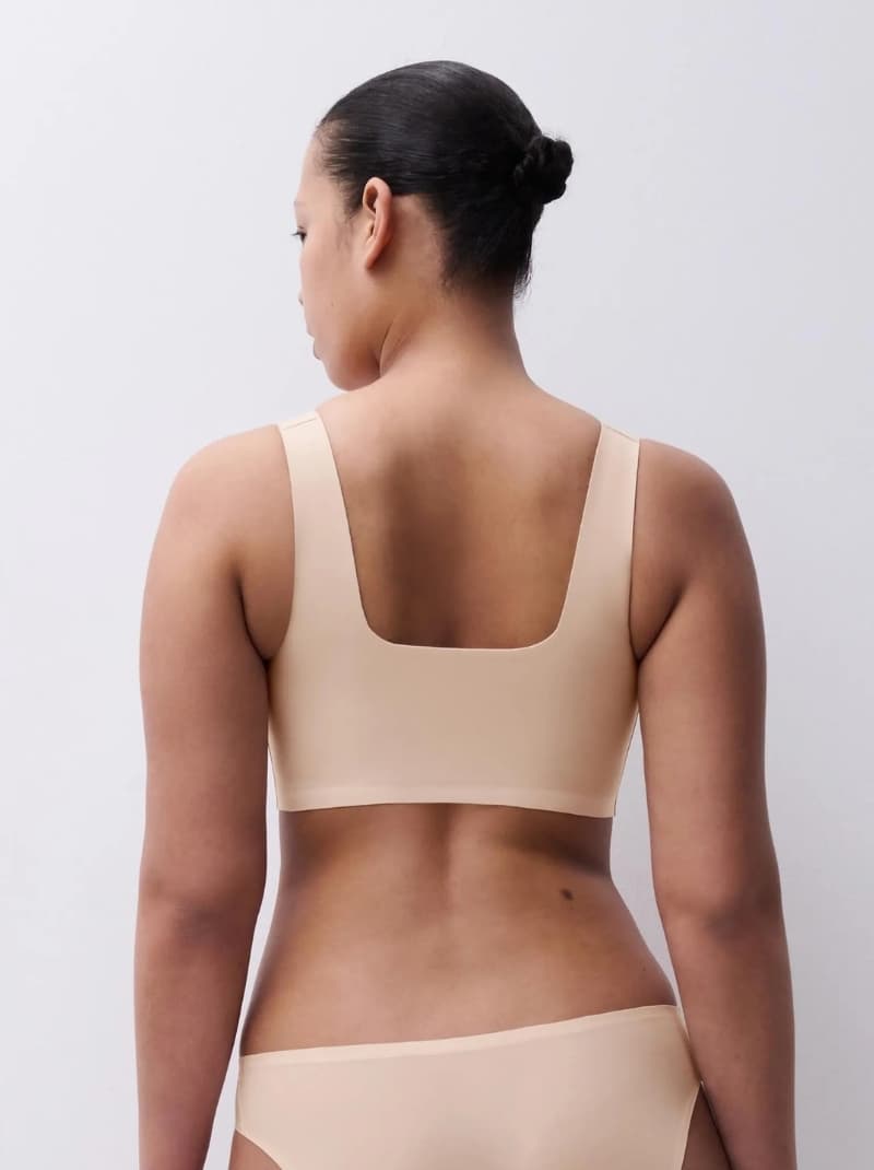 Woman in beige underwear with a squared back neckline, hair in a bun, viewed from behind against a light gray background.