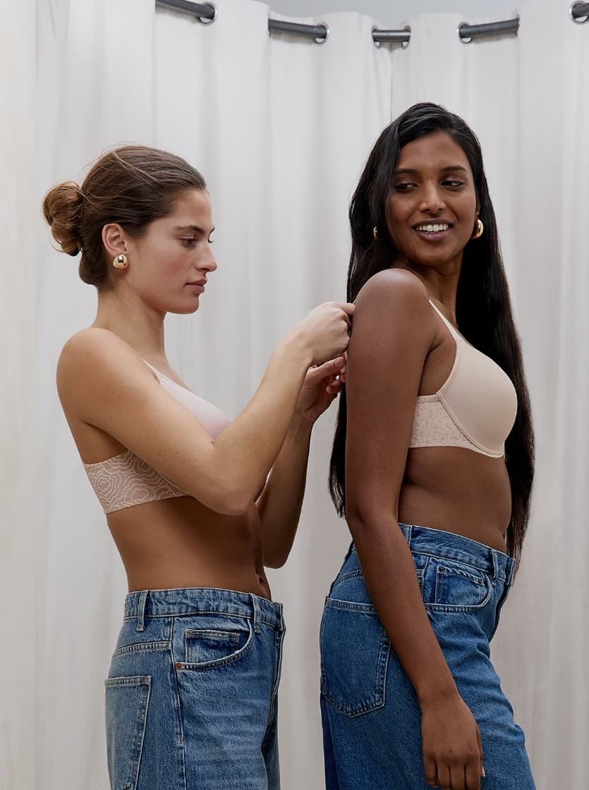 Two women wearing jeans, one adjusting the bra strap of the other, standing in front of a cream curtain, smiling.