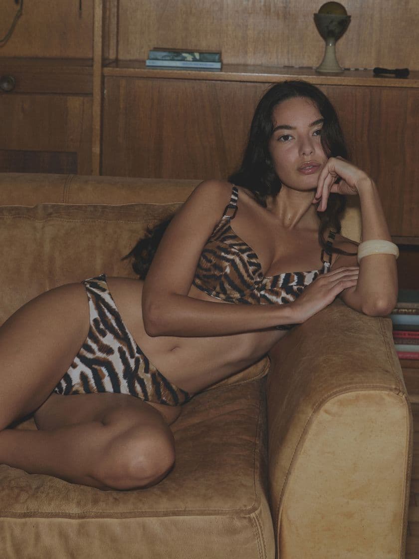 Person in a tiger print bikini lounges on a brown leather sofa, in a room with wooden shelves in the background.