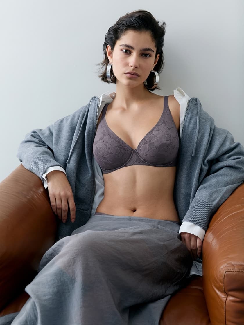 Woman in a grey lace bra and open jacket, seated on a brown leather chair, looking confidently at the camera.