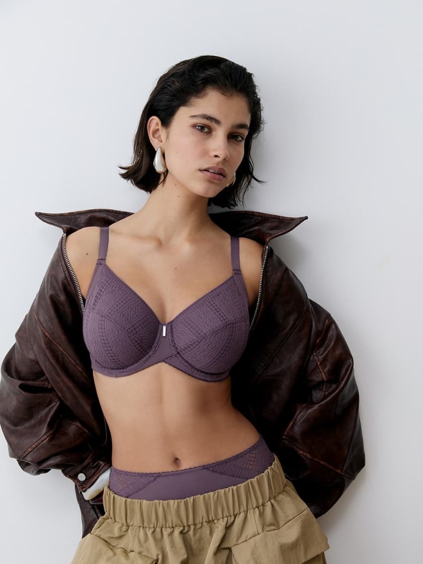 Woman in a purple lace bra, khaki ruffled bottoms, and brown leather jacket, standing against a white background.