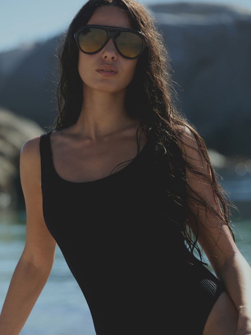 Woman with long dark hair wearing a black swimsuit and large sunglasses stands on a beach, with rocks and water in the background.