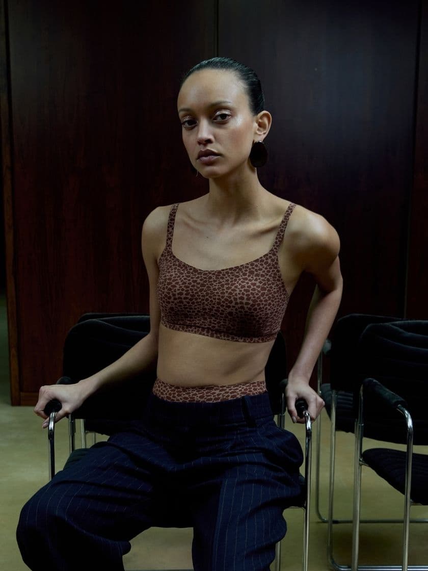 Person seated in a dark room, wearing a leopard-patterned top and striped pants, with a focused expression and hands on chair armrests.