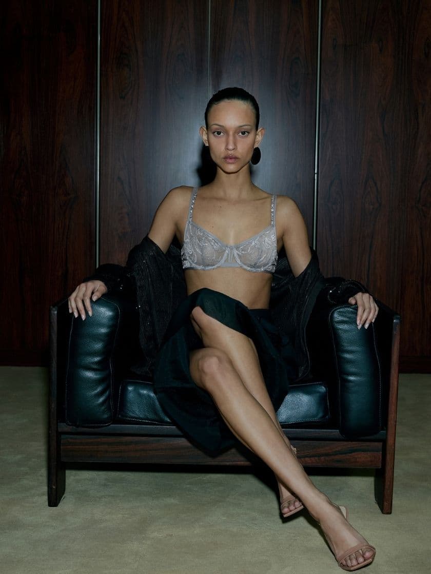 Person seated on a leather chair against a wooden background, wearing a lace bra and draped in a dark garment, with legs crossed and sandals.