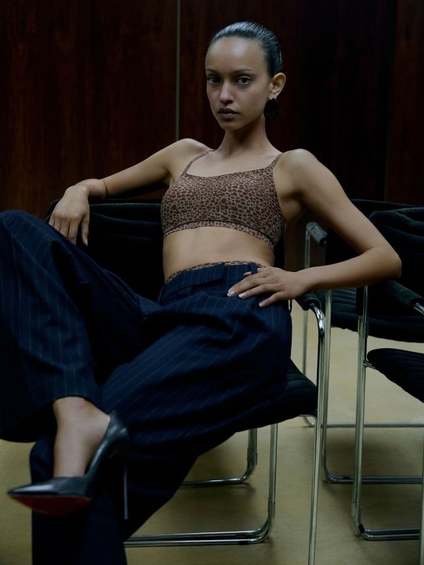 Person sitting confidently in a chair, wearing a leopard print top and pinstripe pants, set against a dark wooden backdrop.