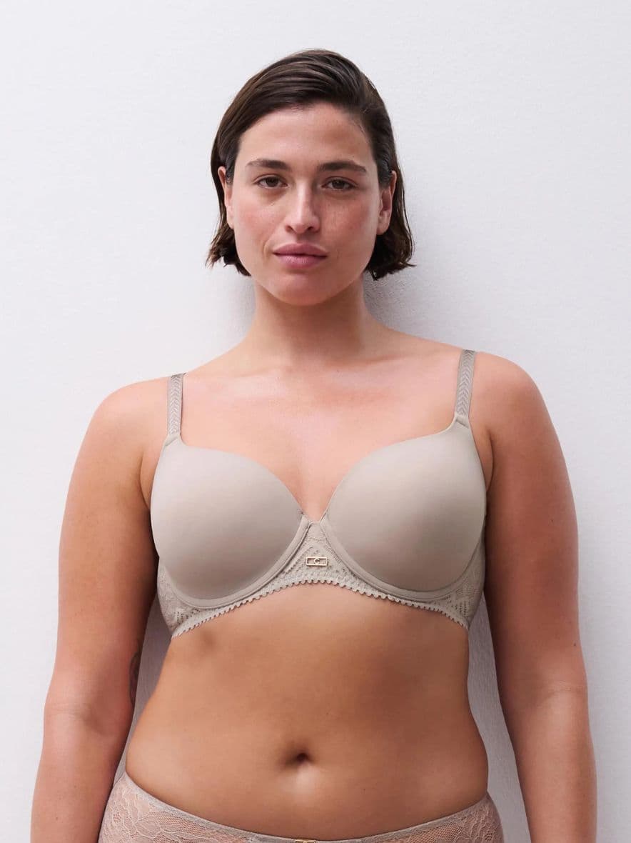 Person wearing a neutral-colored bra stands against a plain white background, looking confidently at the camera.