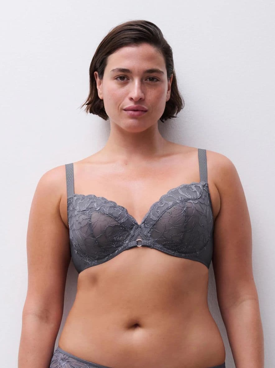 Woman wearing a gray lace bra, standing against a plain white background, looking directly at the camera.