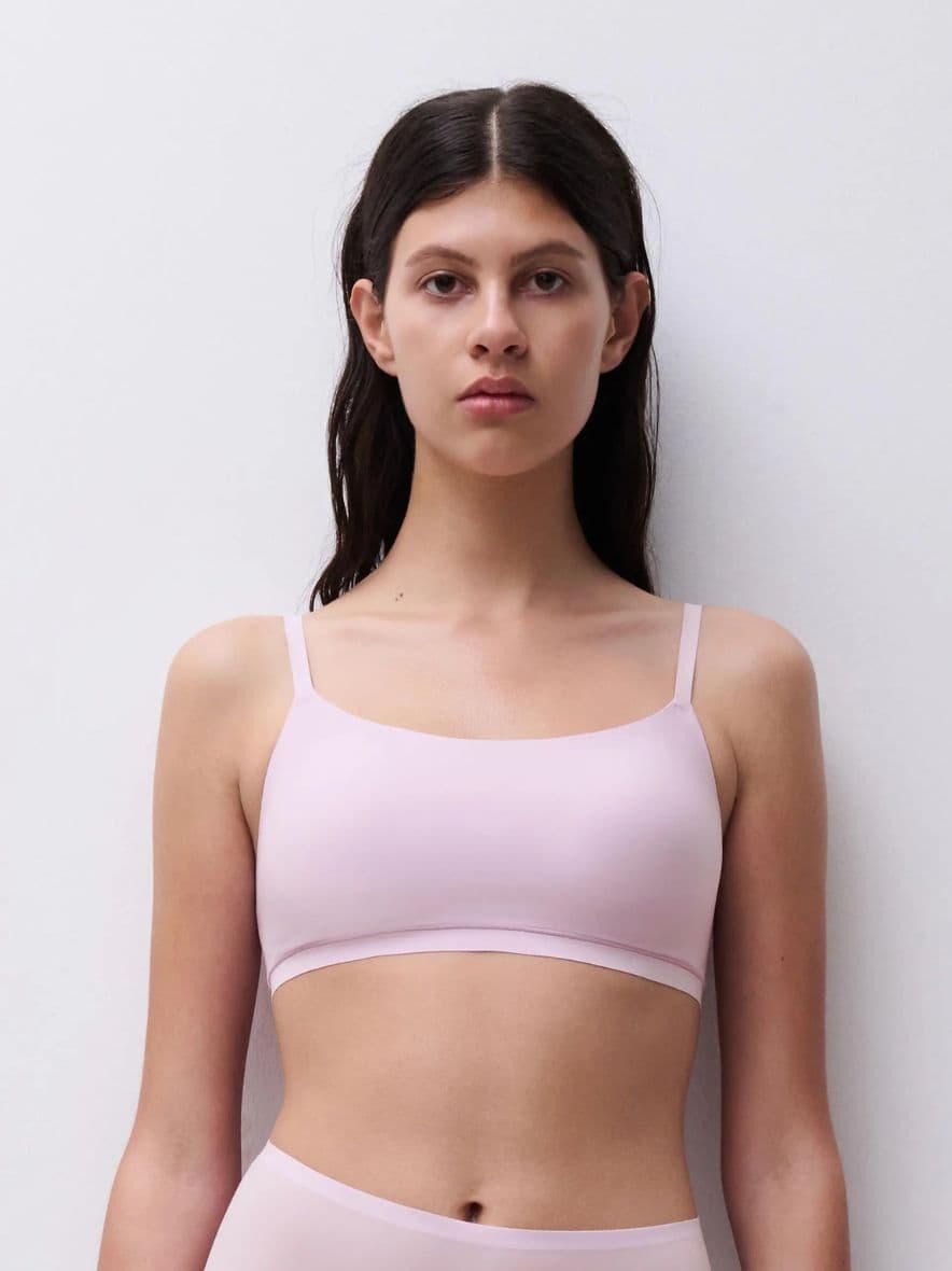 A woman with long dark hair wearing a mauve colored bra stands against a plain white background.
