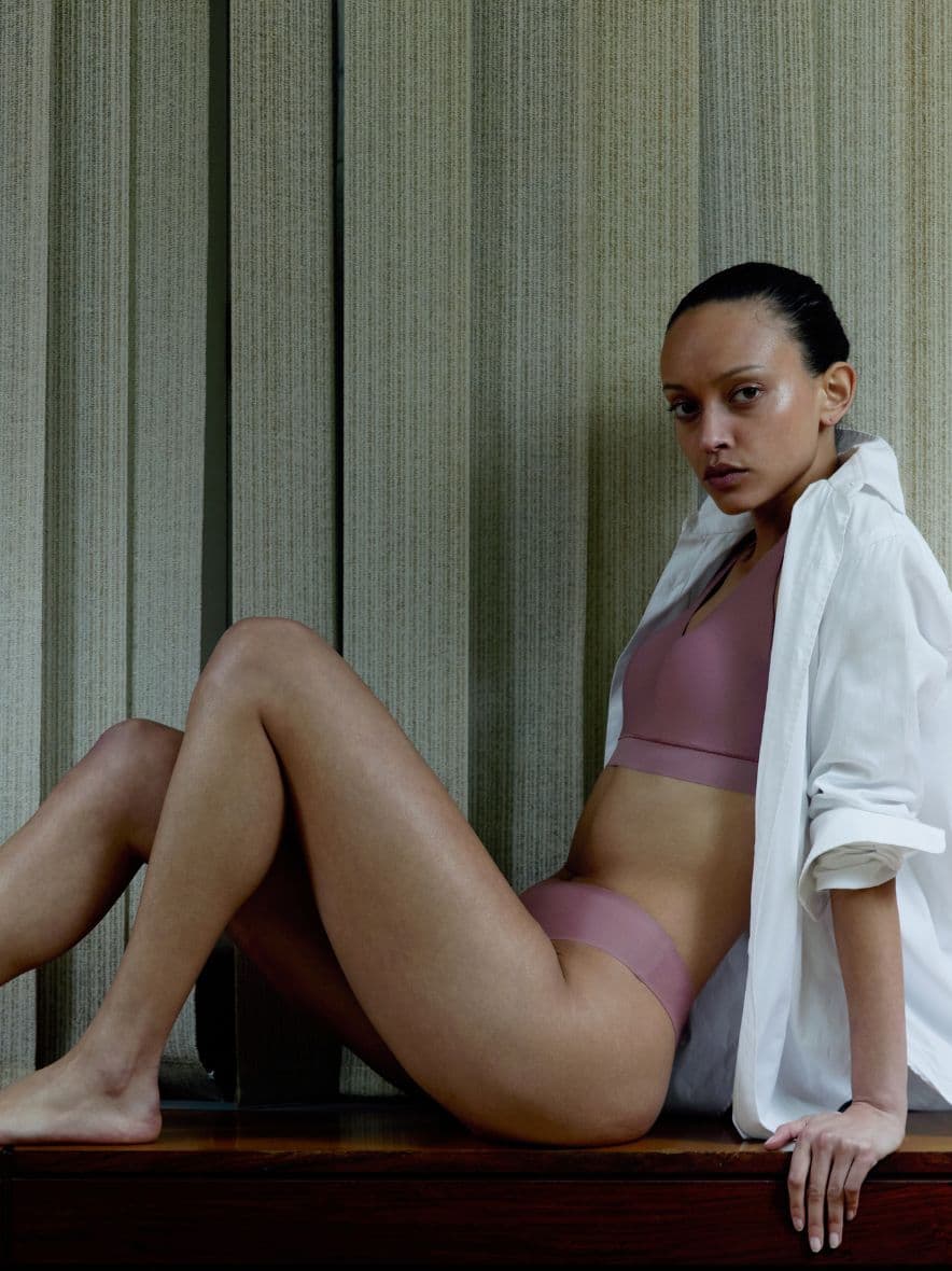 Person in mauve underwear and white shirt sits on a wooden surface, posed confidently against a neutral vertical-paneled background.