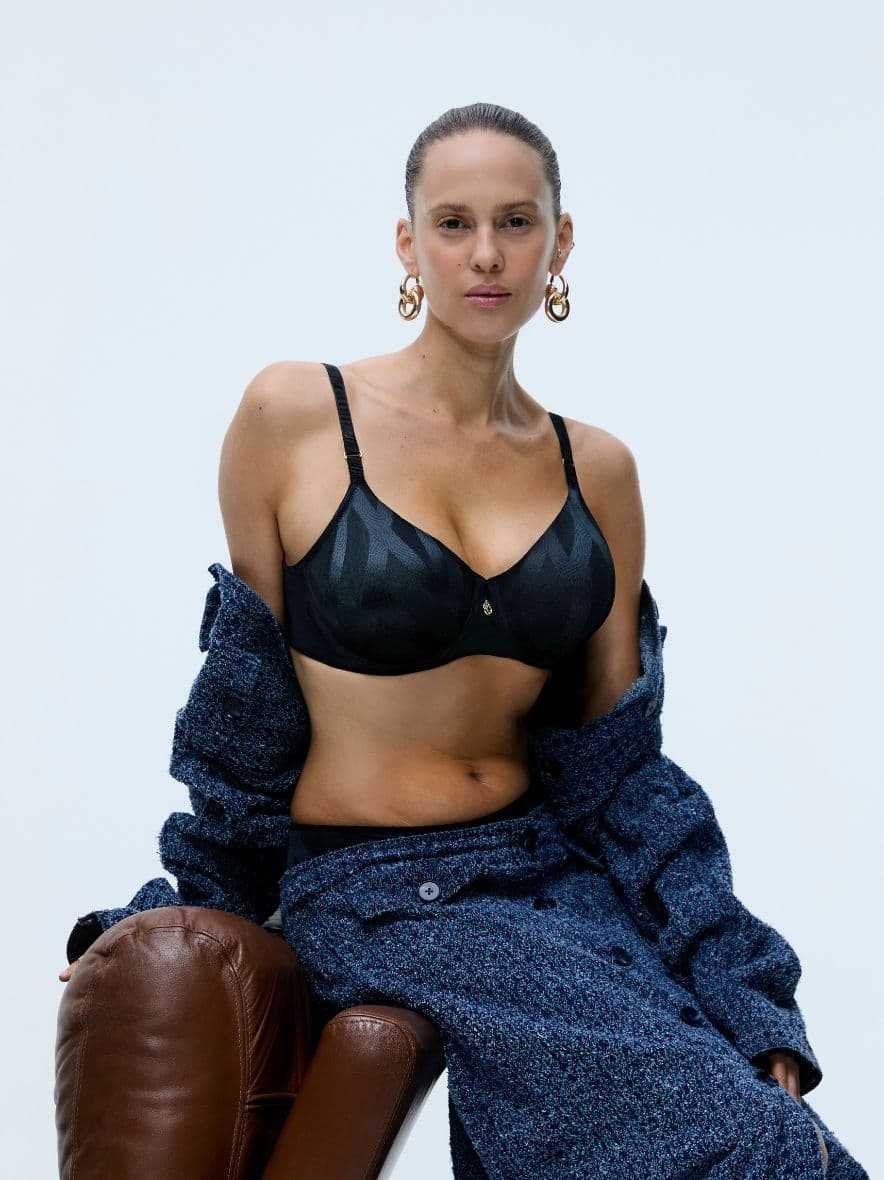 Person posing confidently in a black bra and a blue textured coat, with large hoop earrings, seated on a brown leather surface.