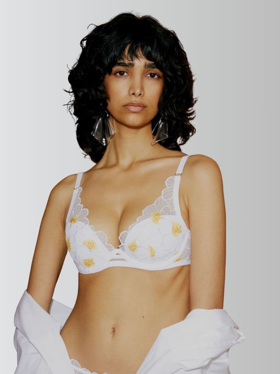 Person with wavy black hair wearing a white floral lingerie set and holding a white shirt off the shoulders, against a gradient background.