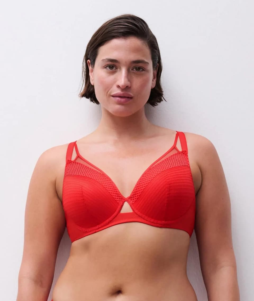 A person with short hair wearing a red bra stands against a plain background, facing the camera.