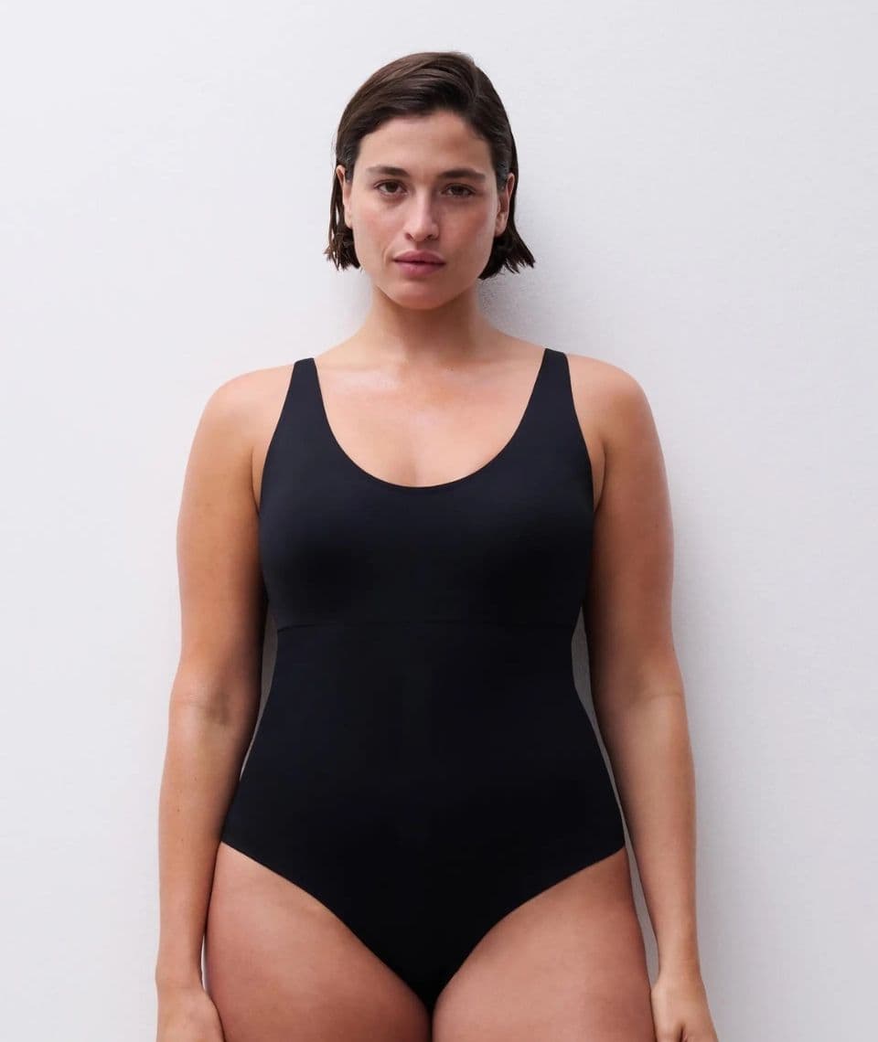 A person in a black shapewear body stands against a plain white background, looking directly at the camera.