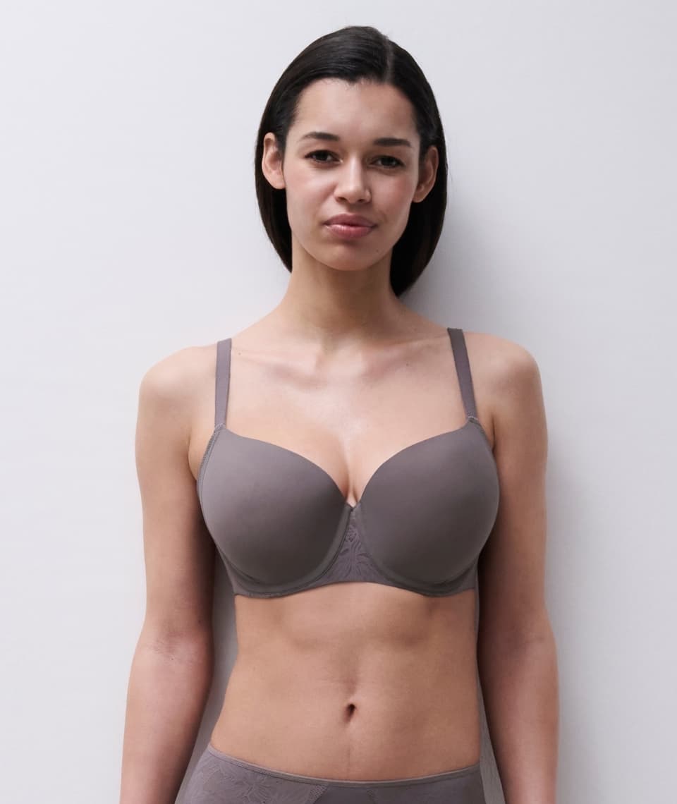 Person wearing a grey bra stands against a plain, light background, looking directly at the camera.
