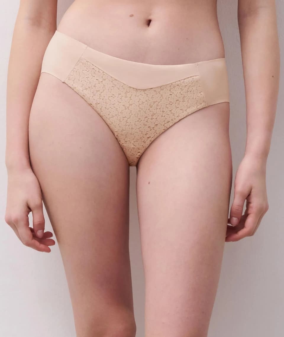 A woman wearing nude-colored lace-patterned high-waisted panties, posing against a plain background.